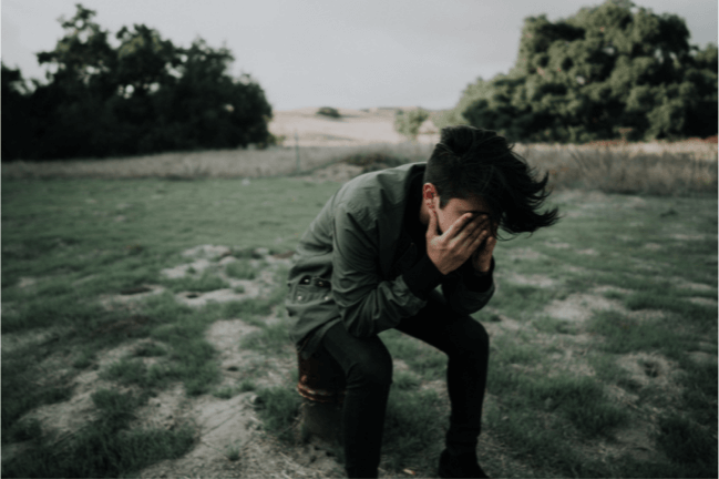 Picture of a man sitting with his palm covering his face on a field with grass and trees behind