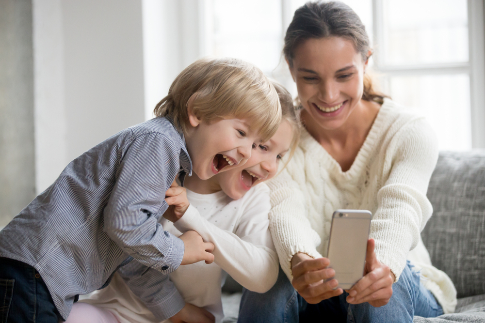 Smiling mother taking selfie with cute kids on smartphone, happy young mom laughing making photo with little son and daughter at home, single mommy and adopted children playing having fun with phone